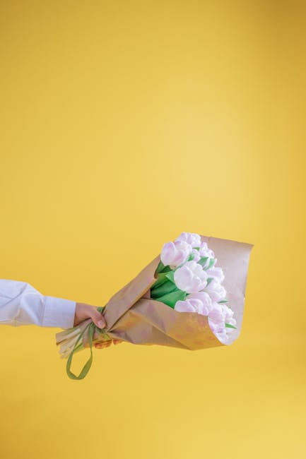 A person's hand, dressed in a white sleeve, holds a bouquet of fresh pink tulips wrapped in light brown paper with a green ribbon, positioned against a bright yellow background. The tulips have smooth