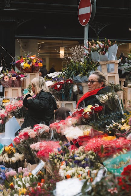 A lively flower market scene in Kilburn, with multiple floral arrangements and bouquets displayed on wooden crates and tables under a dark canopy. The flowers include colorful roses, tulips, daisies,