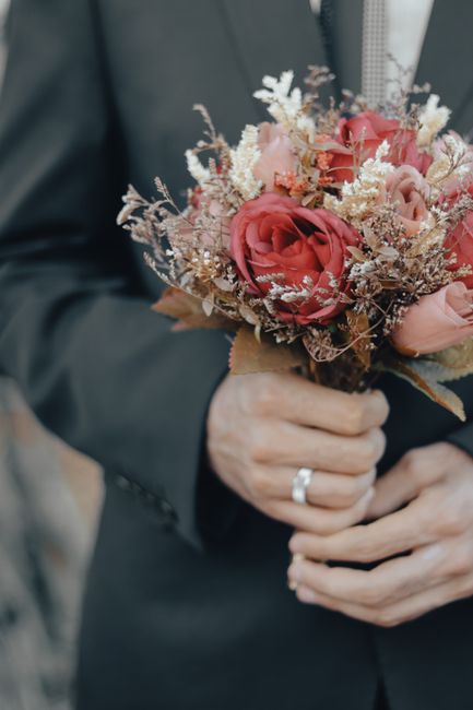 A person dressed in a dark suit is holding a bouquet of fresh flowers. The bouquet features large, velvety pink roses, complemented by smaller white and beige accent flowers with delicate, feathery te
