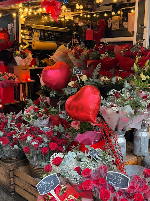 A vibrant display of floral arrangements at a flower shop, featuring predominantly red flowers such as roses, carnations, and other blooms, complemented by white accents and lush greenery. The flowers