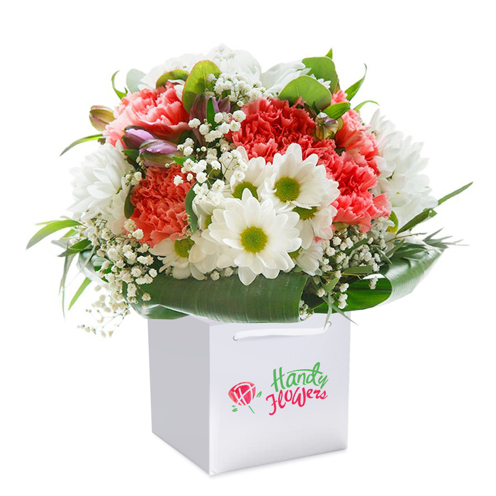Expertly presented in a smooth, square white gift bag, this radiant bouquet is a showcase of Kilburn's floral elegance and local pride. Soft coral and peach carnations dominate with abundant, frilled petals, exuding both texture and a sense of gentle excitement. Threaded throughout the arrangement are pristine white daisies, their chartreuse centers gleaming brightly, offering a fresh, optimistic contrast much like the vibrant community spaces along Kilburn High Road. Sprigs of airy baby's breath bring ethereal lightness, as if echoing the energy of new beginnings or fresh starts in the local area. Glossy, deep green leaves encircle the arrangement, forming a natural, supportive base and providing a lush, structured look. Carefully tucked into the left side, subtle purple-tinged buds peak out-each an emblem of the hidden gems one finds exploring Kilburn's neighbourhoods. The bag itself is designed for effortless gifting, with sturdy ribbon handles hinting at easy delivery to homes, offices, or celebrations across NW6. Every component is arranged by skilled hands into a full, rounded silhouette that speaks of abundance, attention to detail, and anticipation of joy. Illuminated against a seamless, immaculate white backdrop, the bouquet's rich colours and textures invite feelings of freshness, reliability, and warmth-just like a thoughtful visit to a trusted local florist. Ideal for birthdays, anniversaries, or simply bringing a smile to someone special in Kilburn, this display offers not just flowers, but a curated moment of delight.